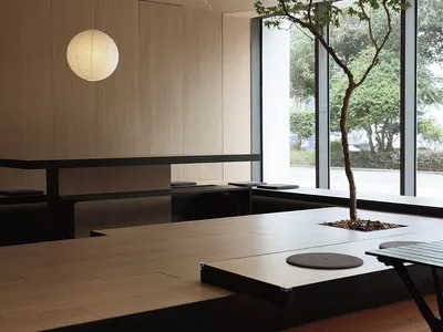 A modern restaurant interior with minimalist design featuring wooden elements and a tree in the center.