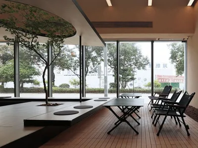 Modern restaurant interior with a wooden floor, tables, and chairs, featuring large windows with trees and greenery visible o