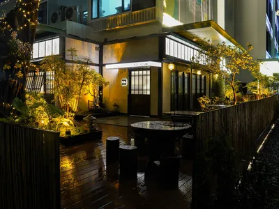 An illuminated, modern Asian restaurant exterior with glass doors and wooden seating areas.