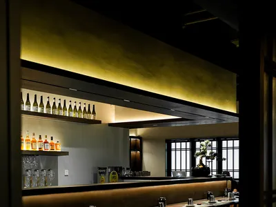 A modern restaurant bar with wooden flooring, tables, and chairs, illuminated by soft lighting.
