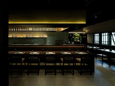 A modern restaurant interior with sleek wooden tables, black chairs, and warm yellow lighting.