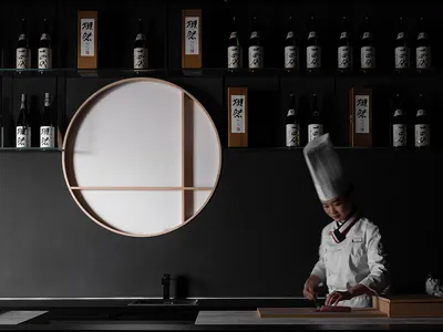 A minimalist restaurant interior with dark walls and a white table, featuring a chef preparing food.