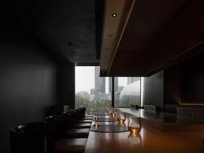 A modern restaurant interior featuring dark concrete walls, wooden tables, and large windows.