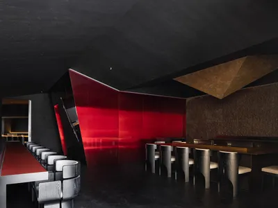 A sleek modern restaurant bar with red leather stools, black concrete walls, and a geometric light fixture.