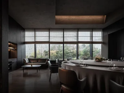 Modern restaurant dining room with round table, chairs, and large windows overlooking greenery.