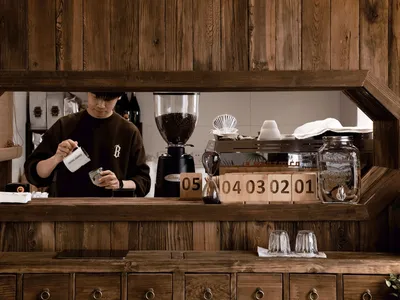 A barista working behind the counter of a coffee shop with wooden materials and a clock showing dates.