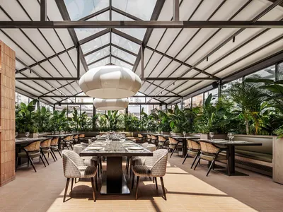 Industrial-style restaurant interior with metal tables and chairs, glass ceiling, and green plants.