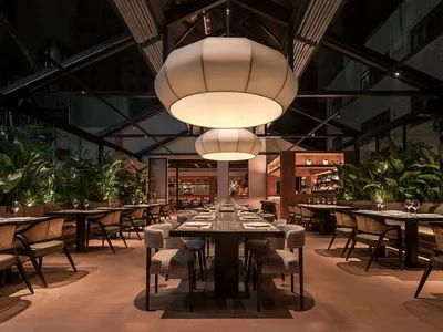 Modern restaurant interior with wooden tables, fabric chairs, and glass pendant lights.