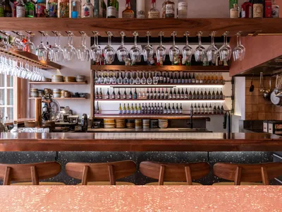 Interior of an industrial-style restaurant bar with wooden furniture and shelves filled with bottles and glasses.
