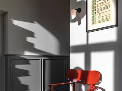A modern minimalist home office with a red chair, certificate frame, and geometric shadows on the wall.
