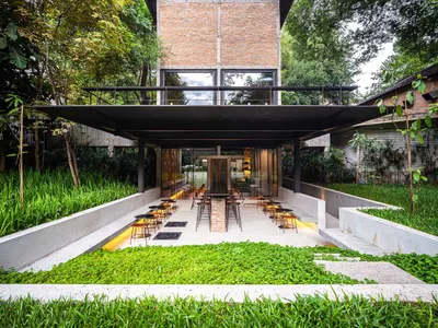 A modern restaurant exterior features a brick façade with a concrete patio surrounded by green plants.