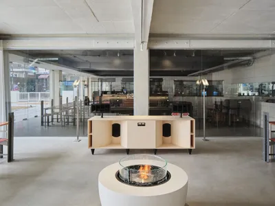 A modern restaurant bar with a concrete floor, industrial-style furniture, and a central fire pit.