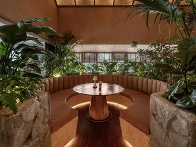 A modern restaurant interior with a circular wooden table surrounded by cushioned seating, concrete walls, and various plants