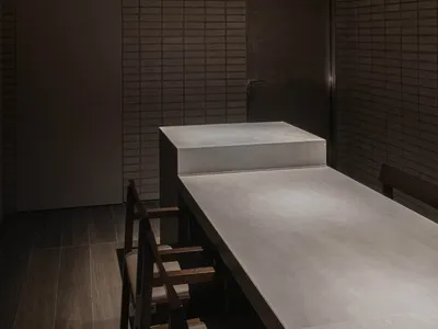 A minimalist restaurant interior with concrete walls, a long table, and dark wooden chairs.