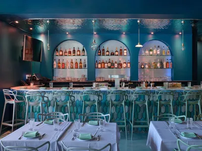 A modern restaurant bar interior with white and green chairs, white tablecloths, and a blue and white tiled counter.