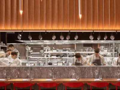 A modern restaurant interior featuring wooden furniture with chefs preparing food in the background.