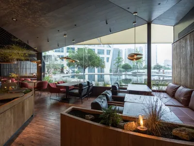 A modern restaurant with wooden furniture, plants, and a view of the city through large windows.