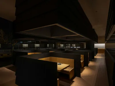 Modern restaurant interior with wooden tables and chairs, concrete walls, and warm lighting.