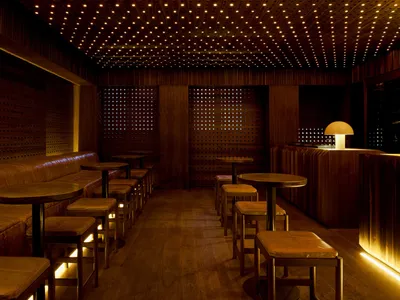 A minimalist restaurant bar with wooden furniture and walls, featuring small round tables and stools.