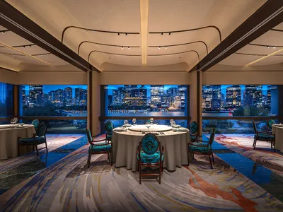A modern restaurant with blue upholstered chairs, glass windows overlooking a cityscape, and a carpeted floor.