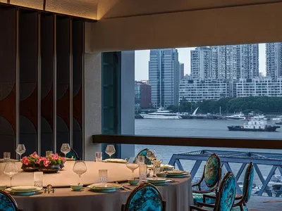 A modern restaurant interior with teal upholstered chairs, round tables, and a view of the cityscape.