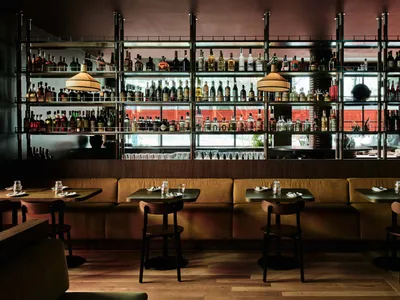 Modern restaurant bar interior with wooden tables, metal stools, and shelves filled with bottles.