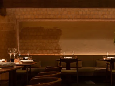 A modern restaurant interior featuring wooden tables and chairs, with a brick wall and green foliage.