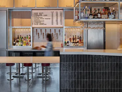 A well-lit, industrial-style restaurant bar counter with a variety of beverages and a menu board hanging above it.