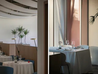 Tables set in a minimalist restaurant interior with white walls and curtains, featuring geometric ceiling design.