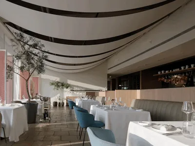 Modern restaurant interior with white tablecloths, blue chairs, white tables, brown flooring, green plants, black vertical st