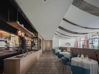 Interior of a modern restaurant with concrete flooring, white tables, blue chairs, and a sleek kitchen counter.