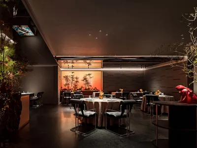 A modern restaurant interior with industrial chairs, tables with tablecloths, and decorative plants.