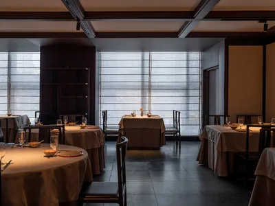 A modern restaurant interior with round tables, chairs, and upholstered seating arranged in a spacious, open layout.