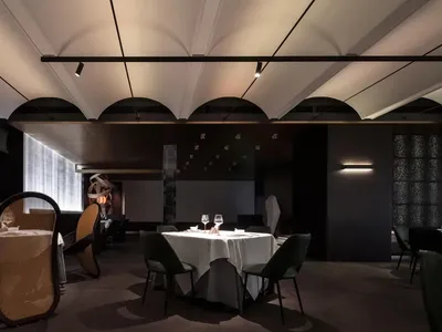Modern restaurant interior with geometric arched ceilings, dark walls, and white tablecloths on tables.