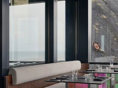 Modern restaurant interior with metal tables, fabric booth seating, and glass elements.