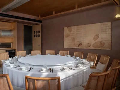Circular table with white tablecloth surrounded by wooden chairs in a modern restaurant interior with concrete walls and larg