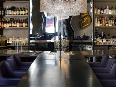 A modern bar counter with a metal finish, featuring a large pendant light fixture and a sleek, minimalist design.