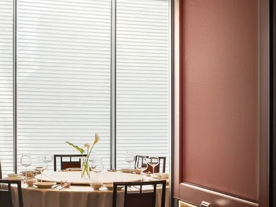 A minimalist restaurant interior featuring white blinds, wooden chairs, fabric tablecloth, and glass drinkware.