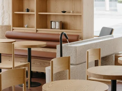 A modern cafe interior featuring wooden tables, chairs, and shelves, along with marble accents and a natural stone seating ar