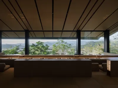 Modern restaurant interior with wooden tables, concrete floor, and floor-to-ceiling windows overlooking natural scenery.