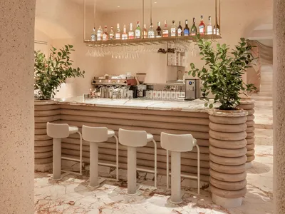 A modern bar interior with marble and concrete materials, featuring a counter, stools, and a shelf with various bottles and g