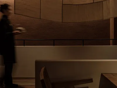 A person wearing a uniform carries a dish in a restaurant setting, featuring modern furniture and wood elements.
