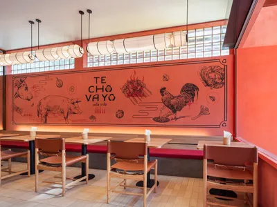 A modern restaurant interior featuring a vibrant red wall mural depicting various food items and the words "TE CHÓ VAYÓ."