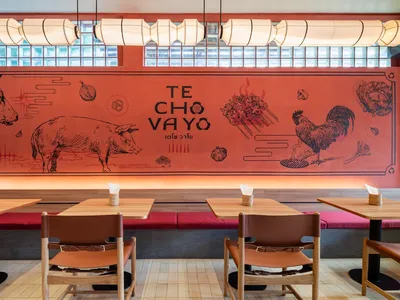 A modern restaurant interior with wooden tables, leather chairs, and a large mural depicting various cooking elements and tex