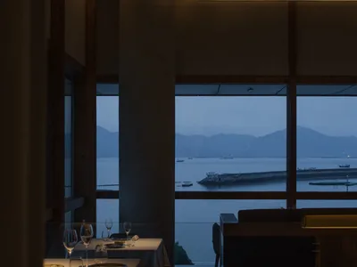 現代簡約餐廳室內，餐桌已擺設晚餐餐具，大型落地窗外可眺望黃昏海景。
