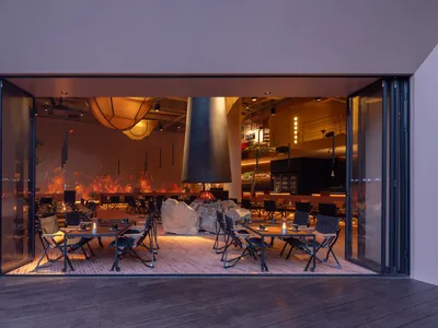 Industrially styled restaurant interior with wooden tables and chairs, a central fireplace, and warm lighting.