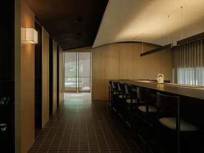 A modern restaurant bar with wooden interior, featuring a long counter and chairs, large windows, and soft lighting.