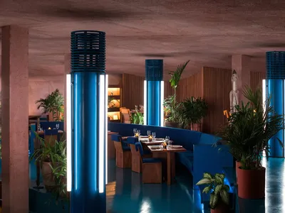 Interior view of a modern restaurant featuring blue light columns, concrete walls, and indoor plants.