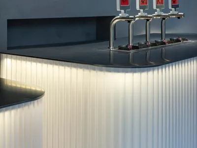 A minimalist restaurant bar with stainless steel fixtures and red signage indicating drink options.