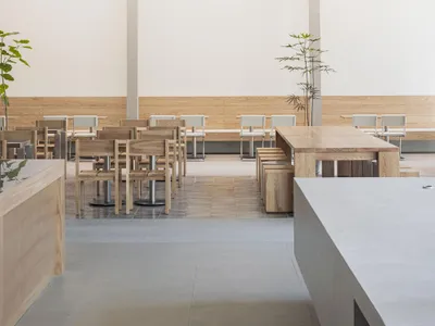 A minimalist restaurant interior featuring wooden tables and chairs with white accents, surrounded by a light-colored wooden
